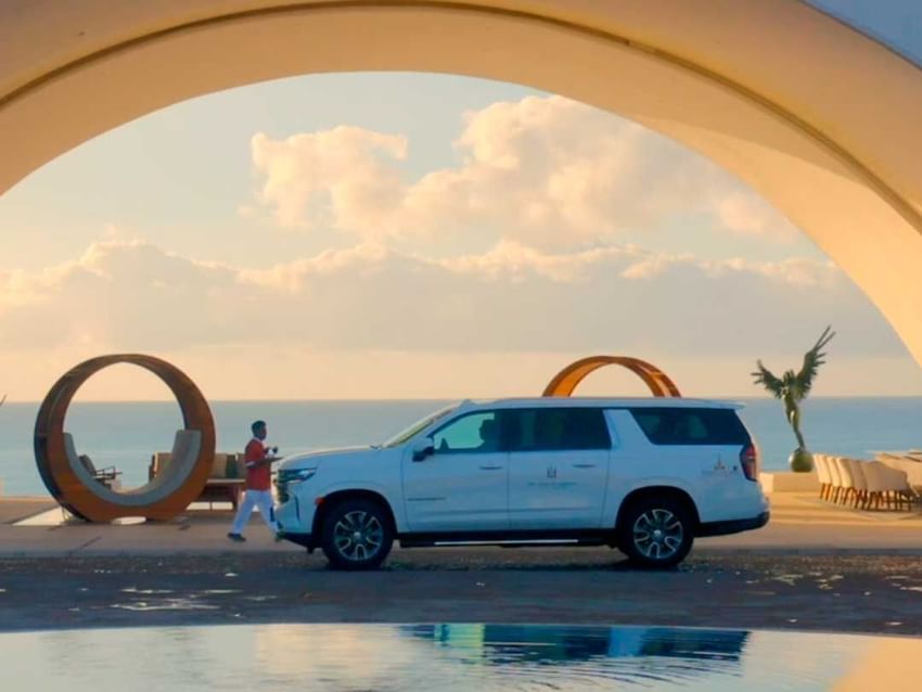A white SUV is parked under an archway at Marquis Los Cabos Resort with the ocean and a person in the background