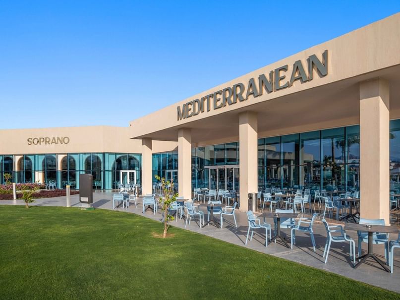 Mediterranean restaurant with outdoor seating, blue tables and chairs, and a green lawn in front.