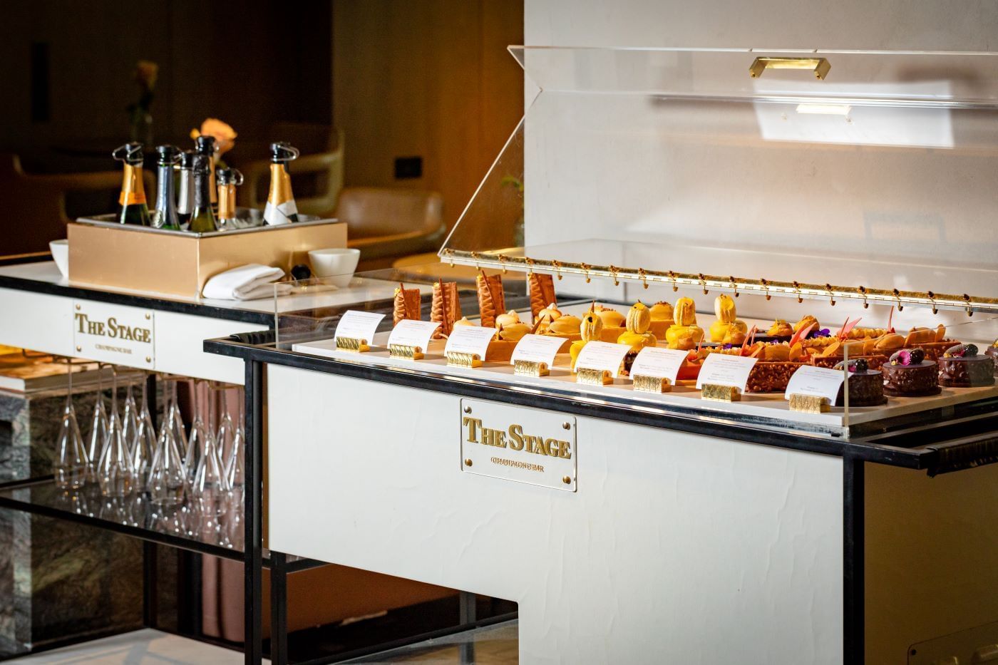 A beautiful display of miniature desserts and champagne bottles on a counter labeled 'The Stage' at The Londoner Hotel