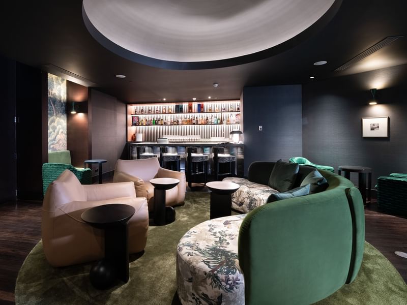 Luxurious bar lounge with green velvet seating, round tables, and a circular rug, featuring a well-stocked bar area.