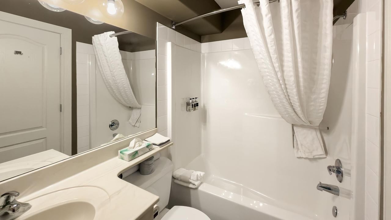 Clean white bathroom with tub, shower curtain, vanity, and mirror at Coast Edson Hotel.
