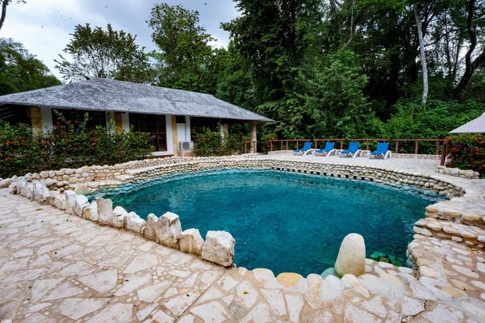 Blue pool by a stone deck with lounge chairs under tropical trees and a grey roof at Hotel Chan-kah Resort Village