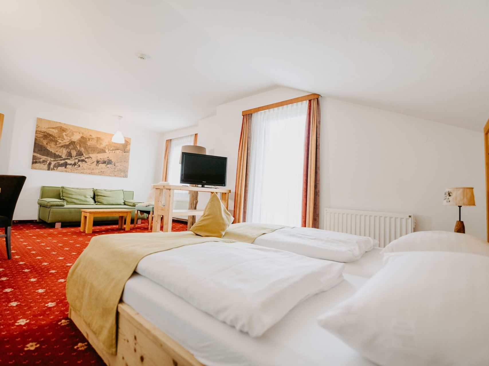 Zimmer mit Doppelbett, rotem Teppich, Sofa, Fernseher und Fenster mit Vorhängen.