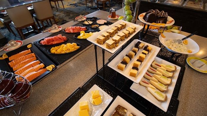 Buffet with fresh sliced fruit and assorted cakes displayed on raised platters in Palmira Cafeteria at Araiza Calafia