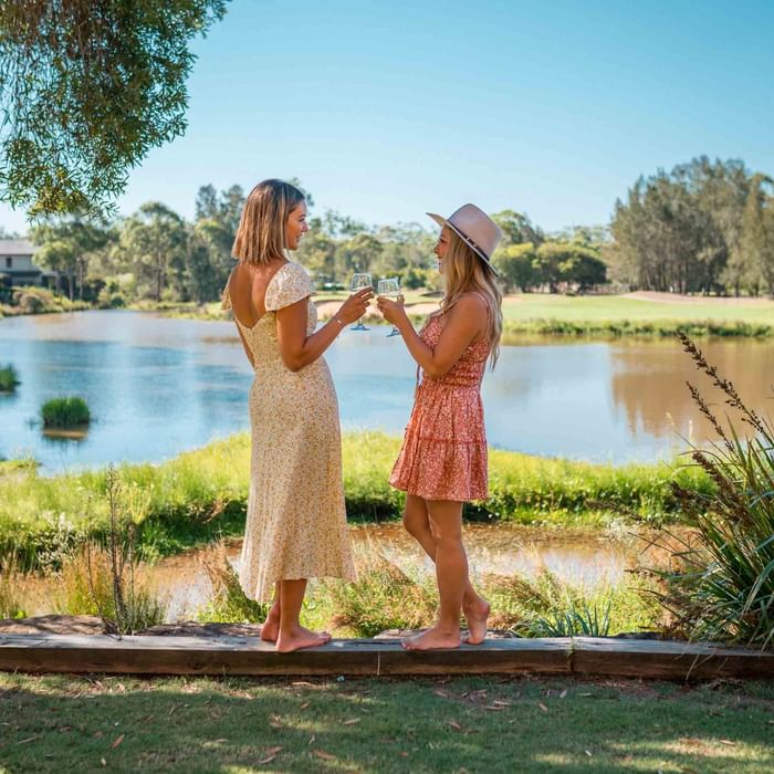 Two women in summer dresses holding wine glasses by a lakeside near Mercure Kooindah Waters
