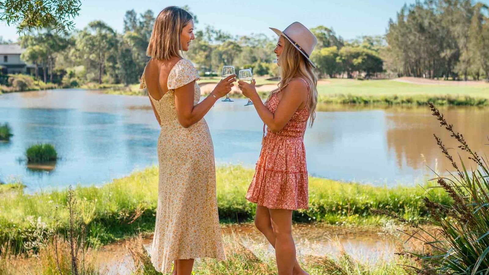 Two women in summer dresses holding wine glasses by a lakeside near Mercure Kooindah Waters