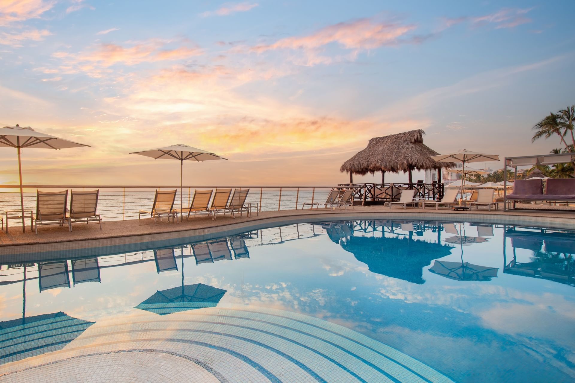 Piscina con tumbonas, sombrillas y un gazebo de techo de palma durante un atardecer con vistas al mar en el Sunset Plaza Beach Resort