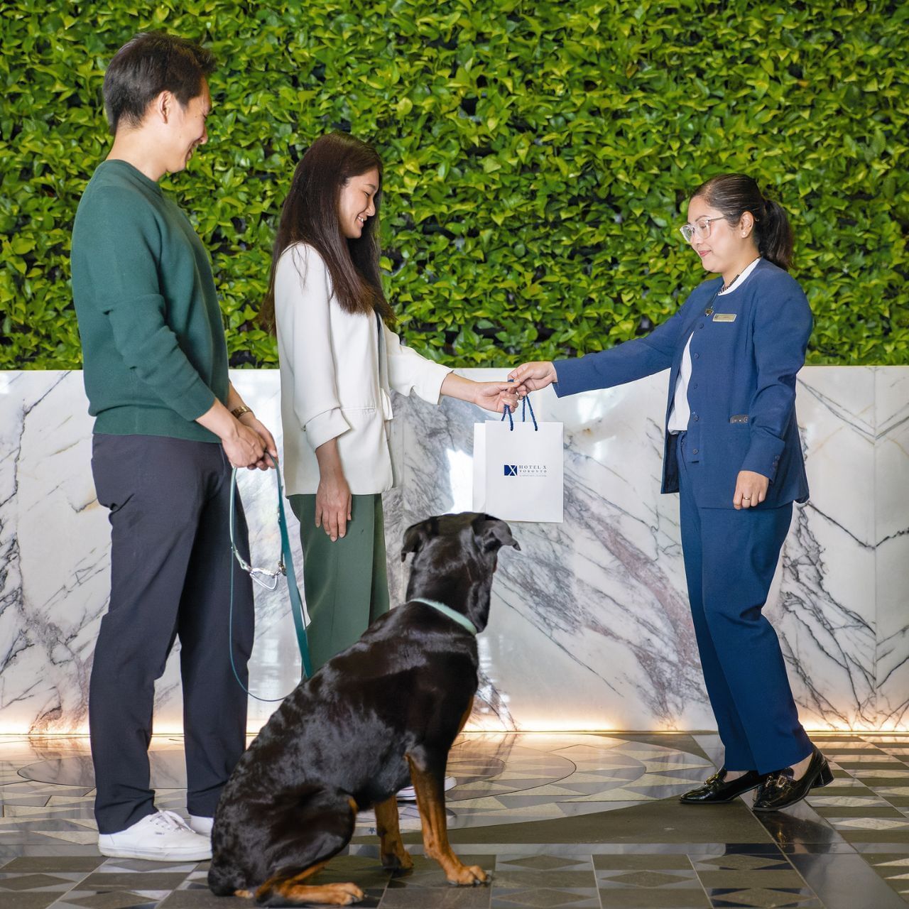 Hotel staff in blue suit handing white bag to couple standing with Rottweiler dog in lobby at Hotel X Toronto