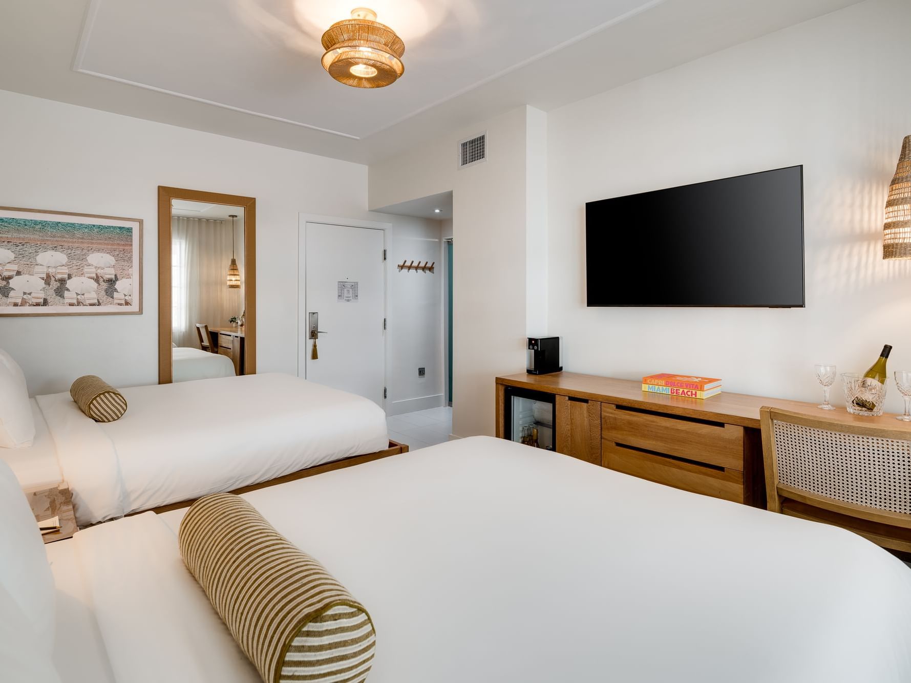 Two beds with pillows, a desk with wine glasses, a TV, and a mirror in a room.