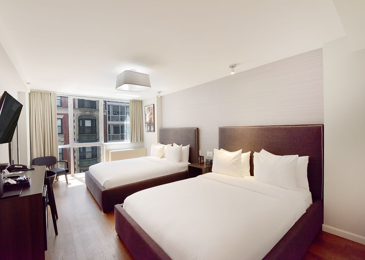 A hotel room with two beds, a desk, a chair, and a window.