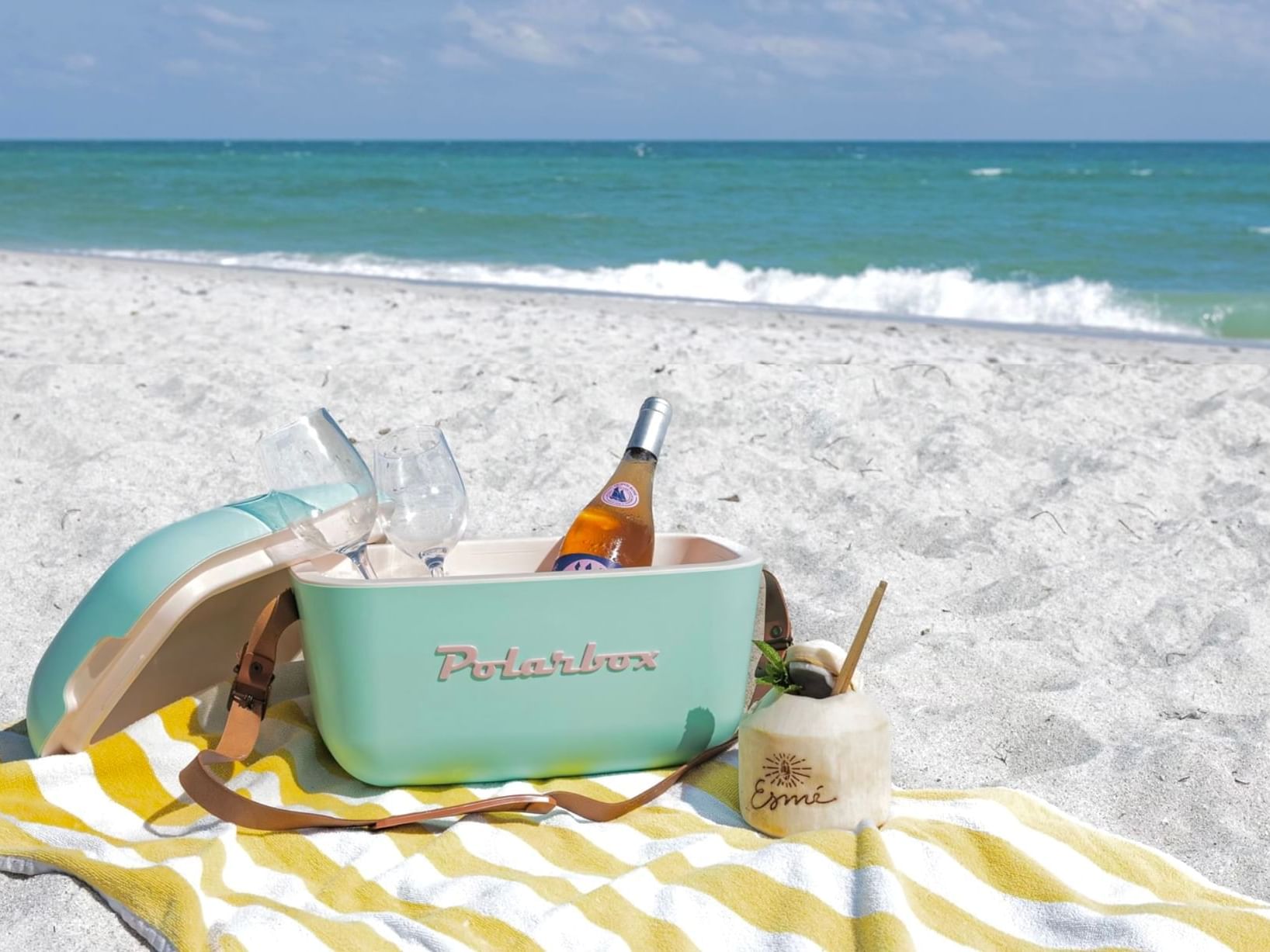 Cooler with a bottle and glasses on a beach blanket by the ocean.