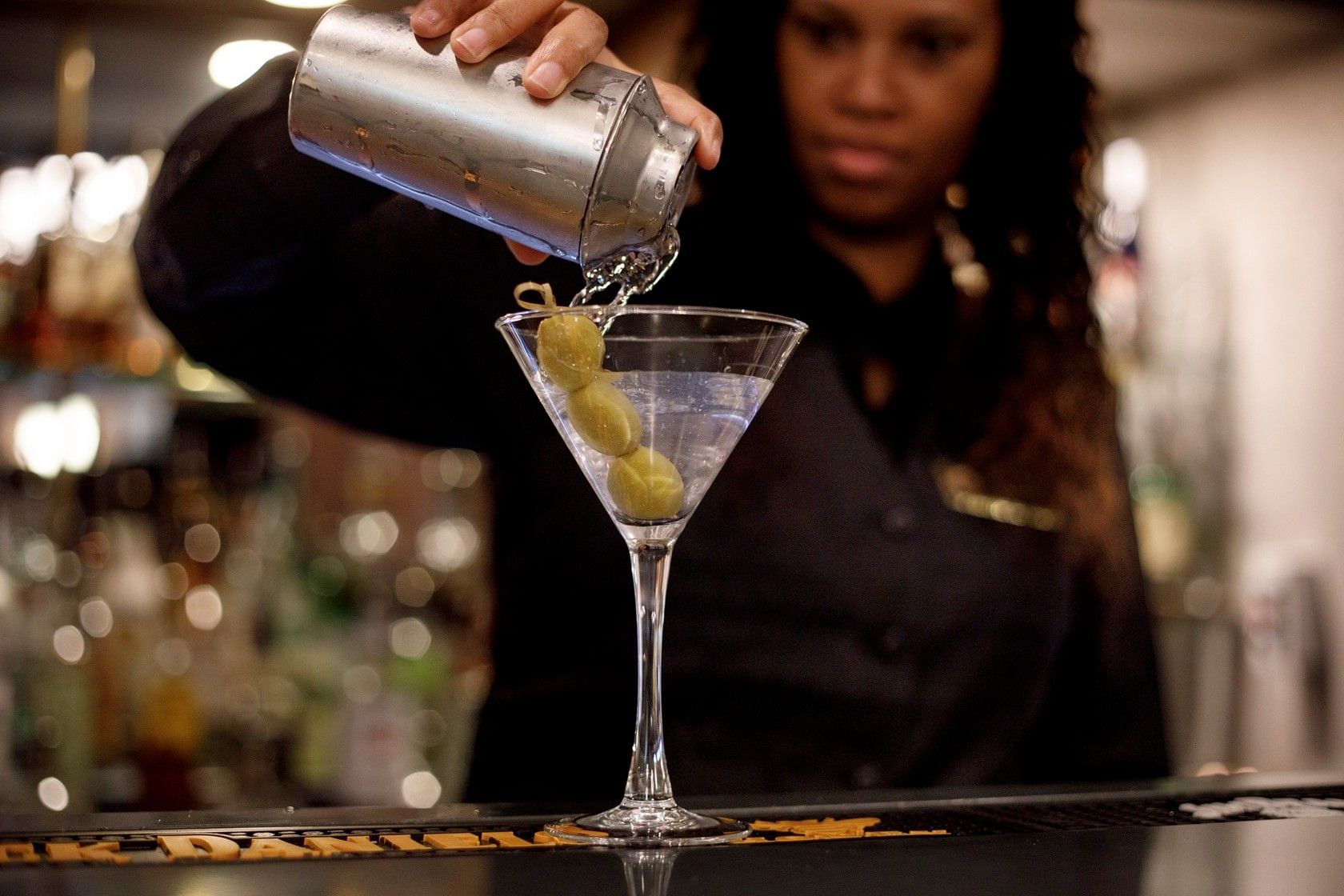 A bartender pouring a drink into a martini glass with olives.