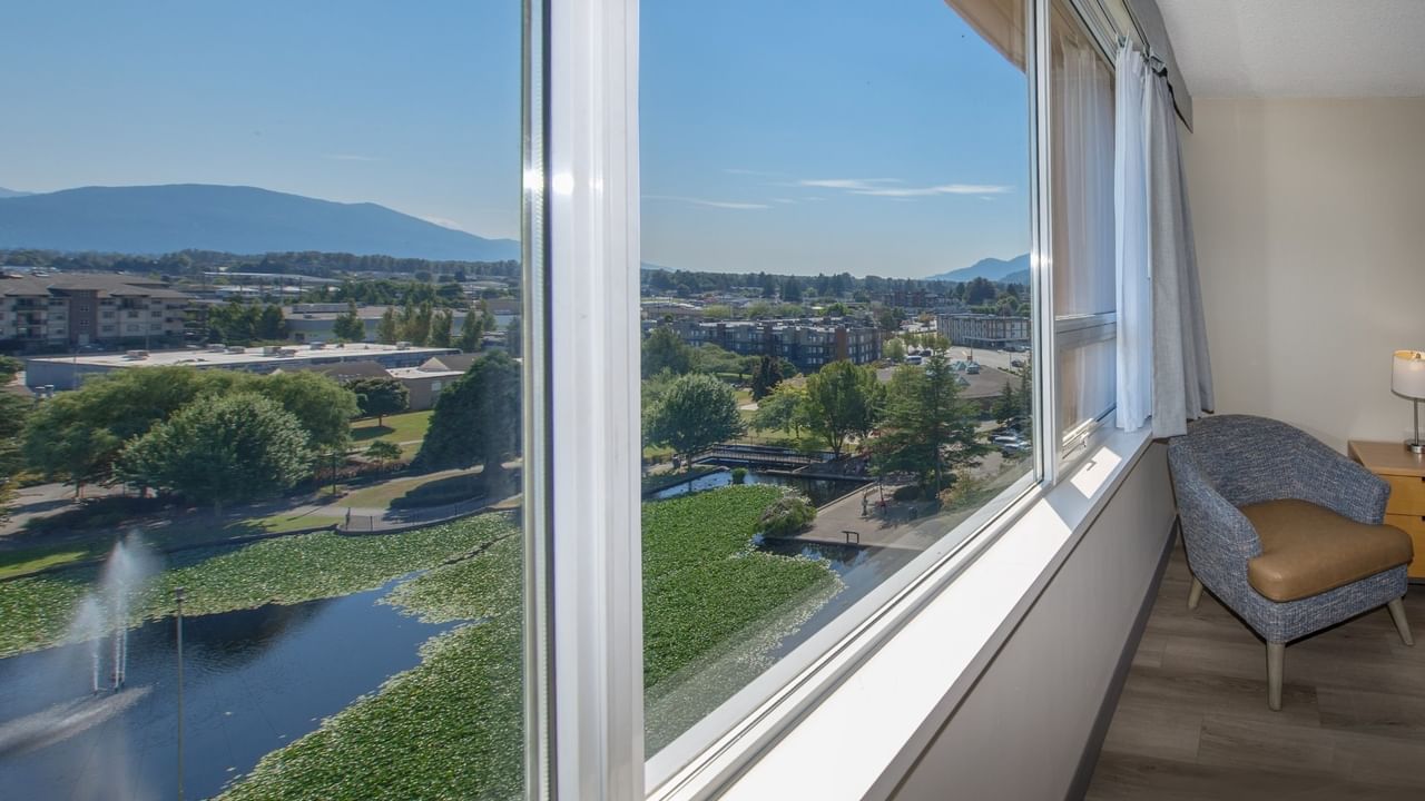 View from a hotel room with large windows showing a pond, cityscape, and mountains.