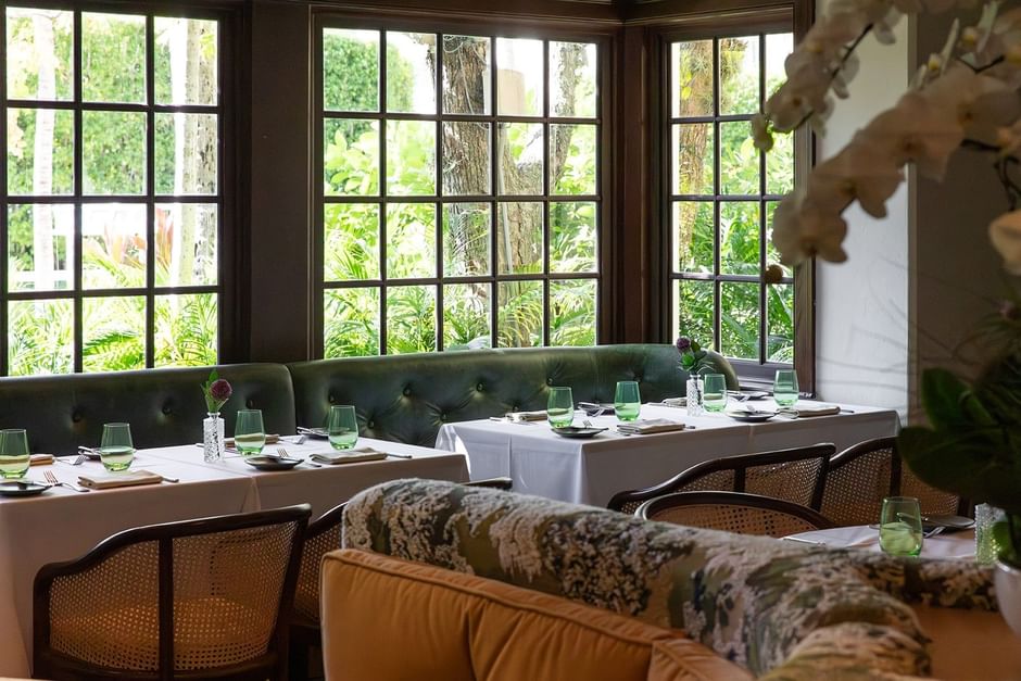 Dining area with green couches, rattan chairs, and tables set with glassware, overlooking greenery through large windows.