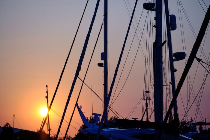 Sailboat masts and rigging silhouetted against a colourful sunset sky with the sun low on the horizon.