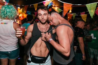 Two men at a festive party are playfully lifting their shirts, showcasing their toned abs at The Gaythering