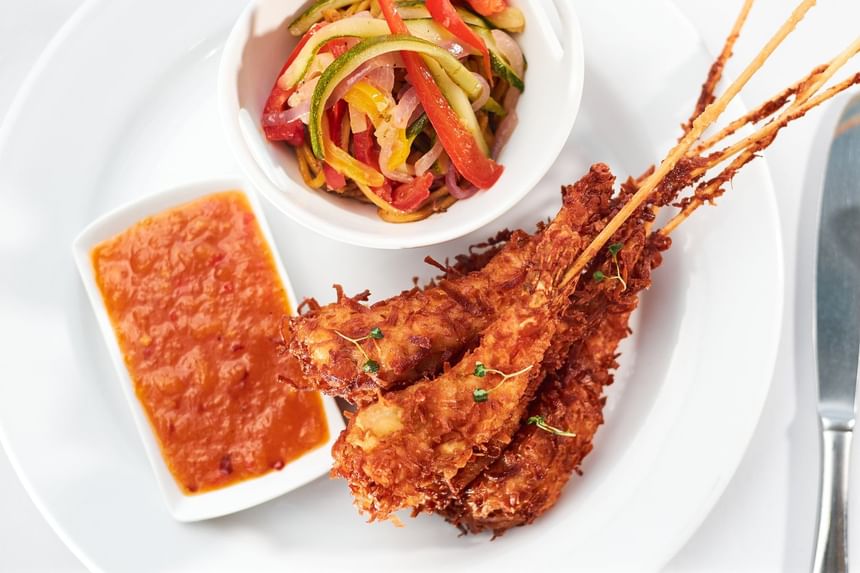 Crispy fried shrimp skewers served with a spicy dipping sauce and a side of colorful sautéed vegetables at Bay Gardens Hotel