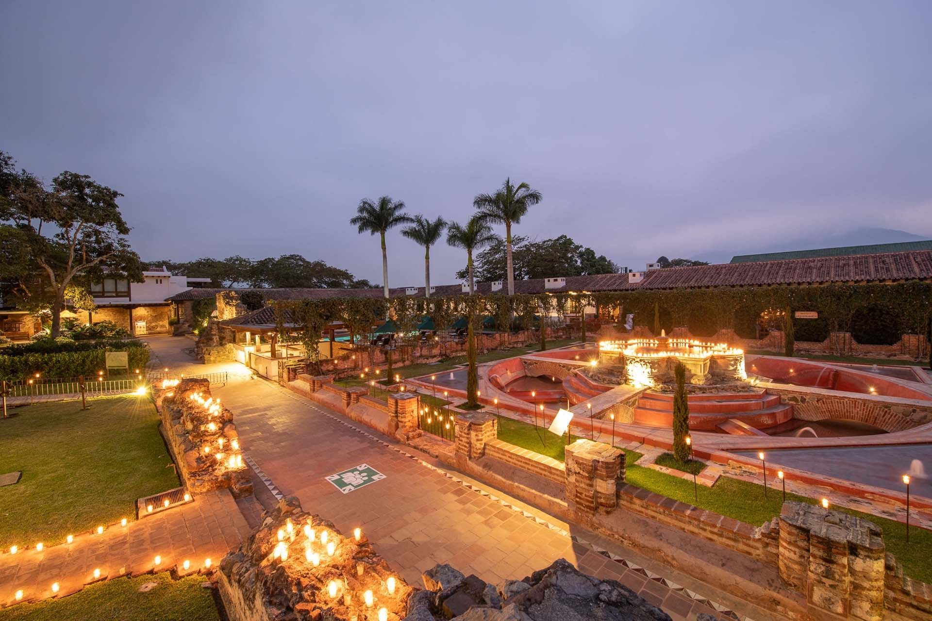 Toma amplia de la fuente iluminada y los terrenos del hotel con palmeras al anochecer en Casa Santo Domingo