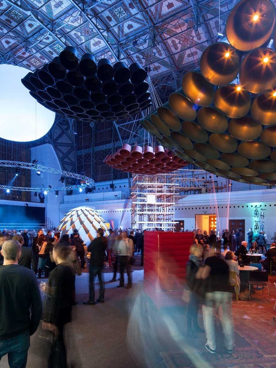 Diverse guests gather under a grand ceiling with artistic lights and hanging art near Almanac X Alcron Prague