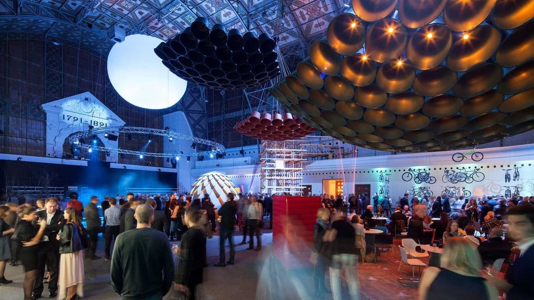 Diverse guests gather under a grand ceiling with artistic lights and hanging art near Almanac X Alcron Prague