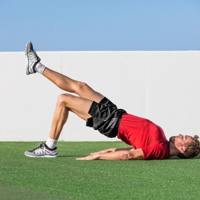 Man performing a single-leg glute bridge exercise outdoors on artificial grass against a white wall and blue sky.