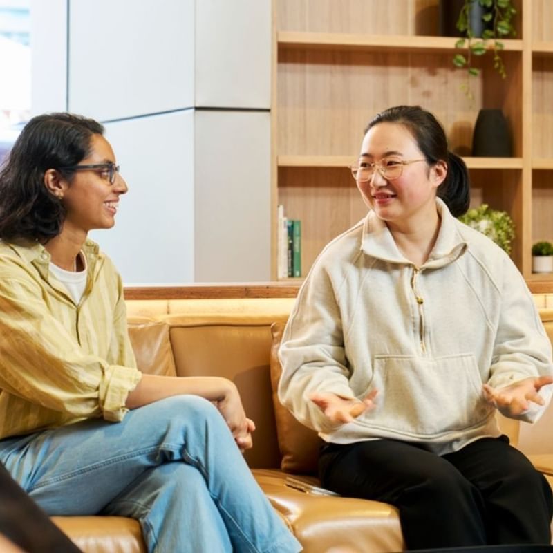 Two students conversing on a tan leather sofa in a bright modern lounge with wooden shelving and plants.