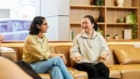 Two students conversing on a tan leather sofa in a bright modern lounge with wooden shelving and plants.