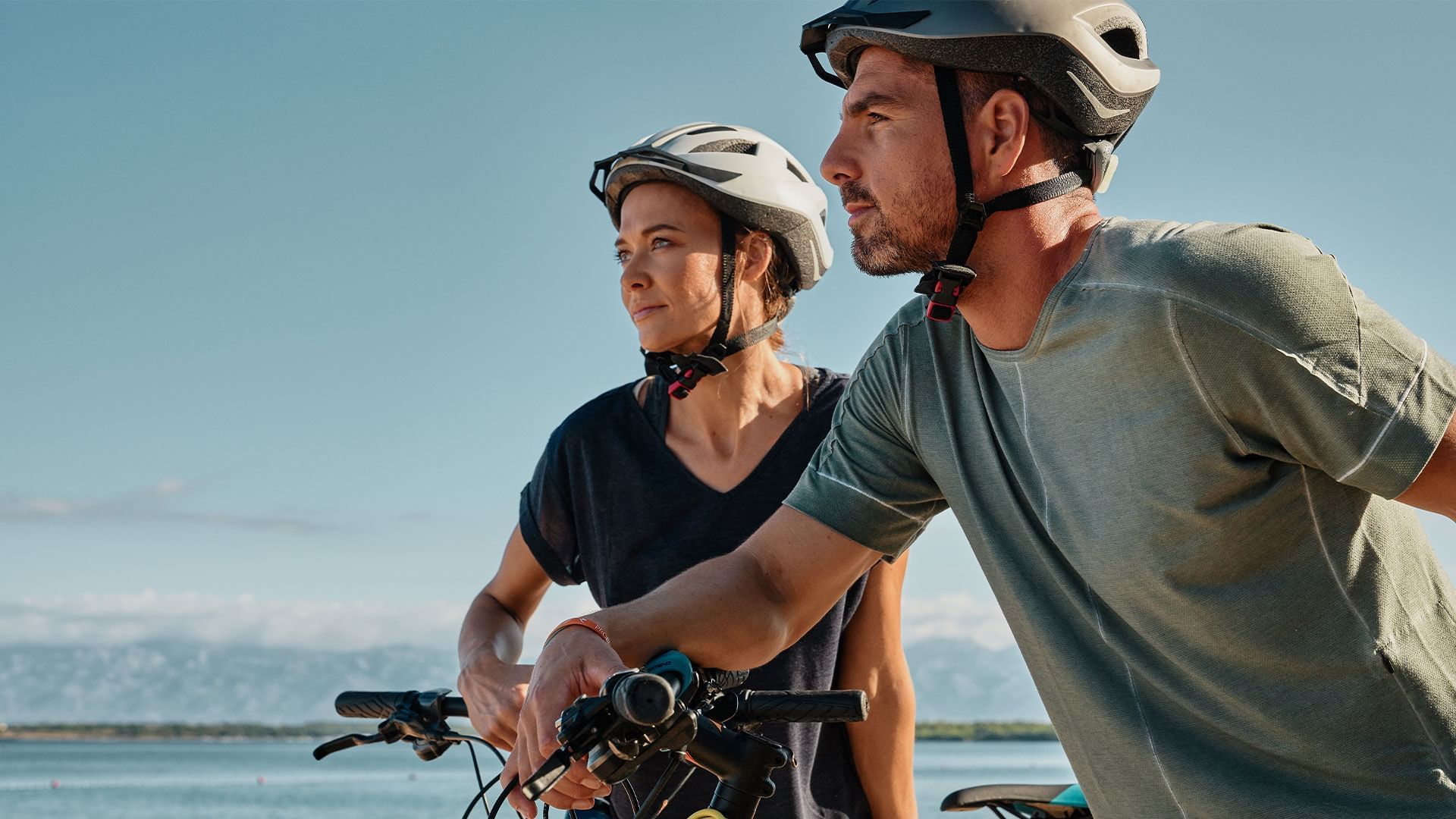 Zwei Radfahrer mit Helmen und sportlicher Kleidung stehen nebeneinander vor einer Wasserlandschaft.