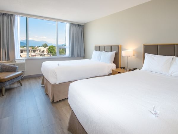 Two beds, nightstands, lamps, and a chair near a window with a mountain view.