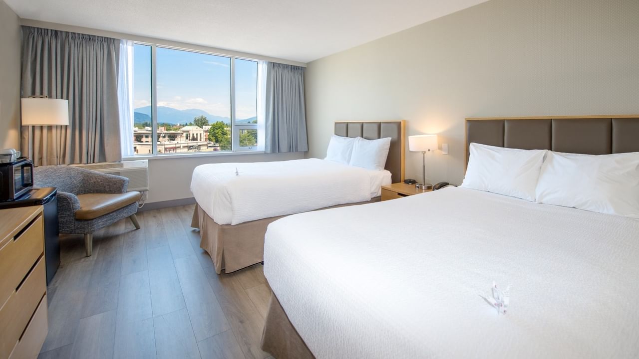 Two beds, nightstands, lamps, and a chair near a window with a mountain view.