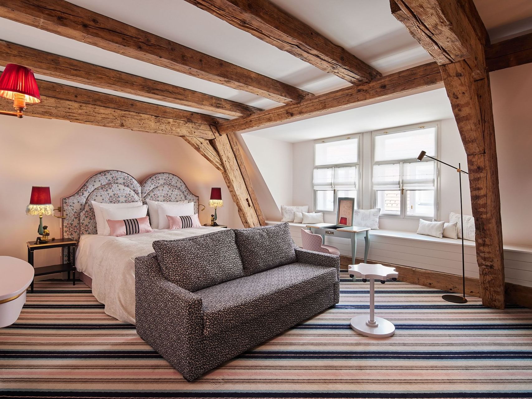Cozy hotel bedroom with exposed wooden beams, striped rug, large bed, sofa, side tables with lamps, and window bench.