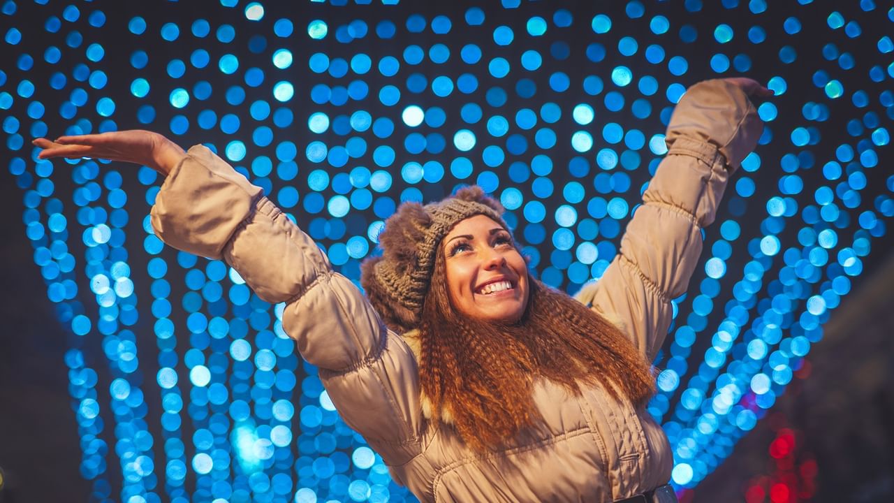 Woman with arms raised joyfully in front of illuminated blue heart display at Winter Lights Festival.