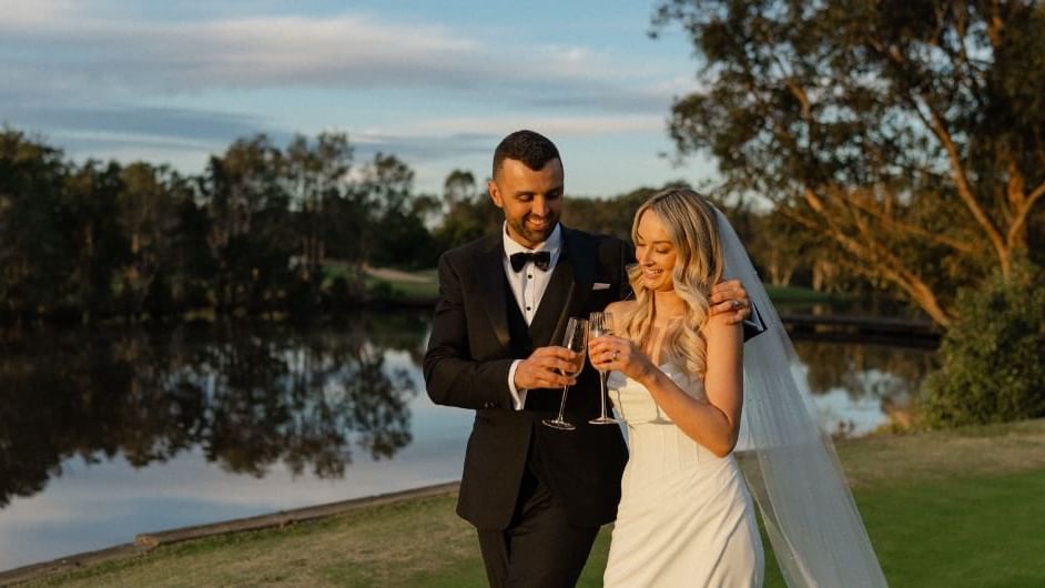 A couple in wedding attire toasting with champagne by a lakeside near Mercure Kooindah Waters