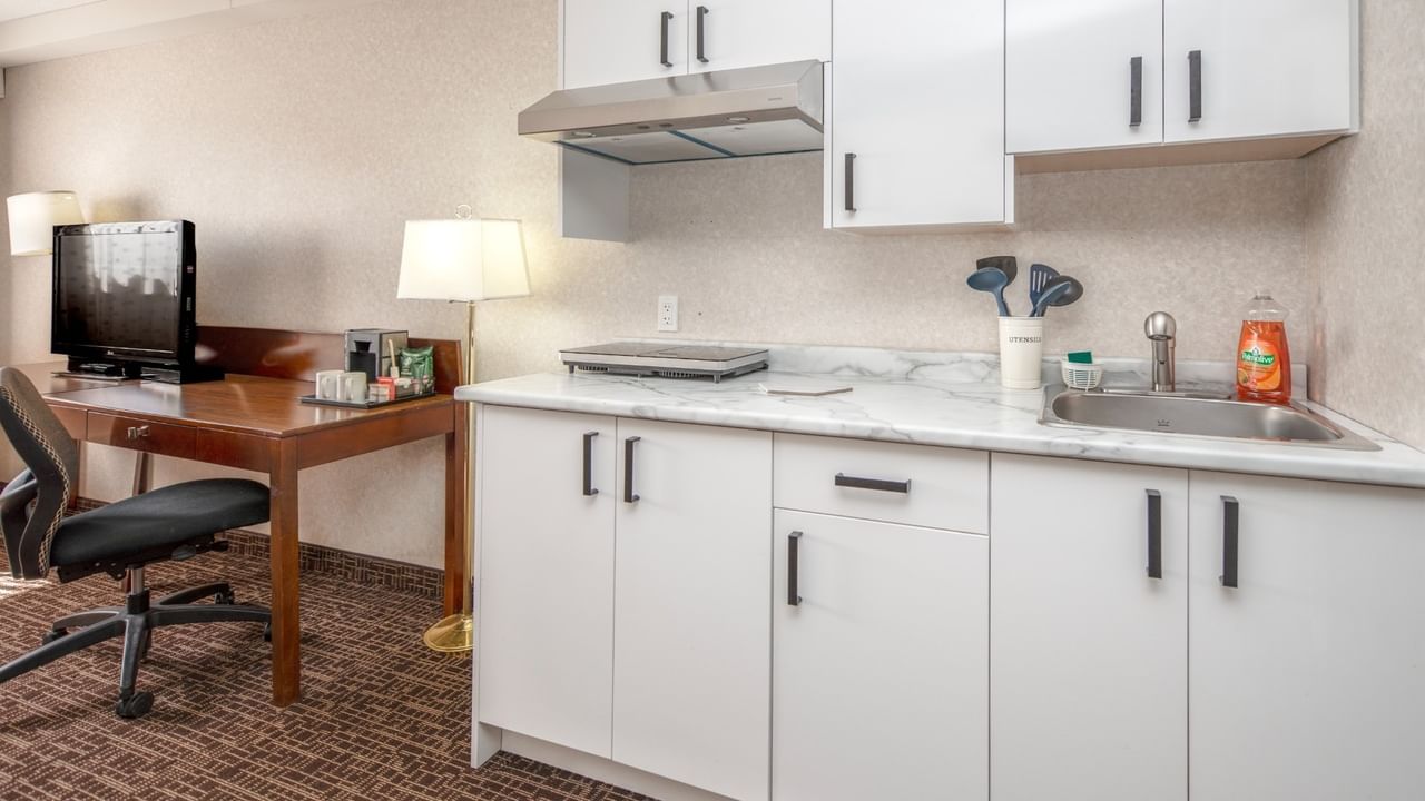 Hotel kitchenette with white cabinets, marble countertop, sink, and adjacent desk with TV and chair.