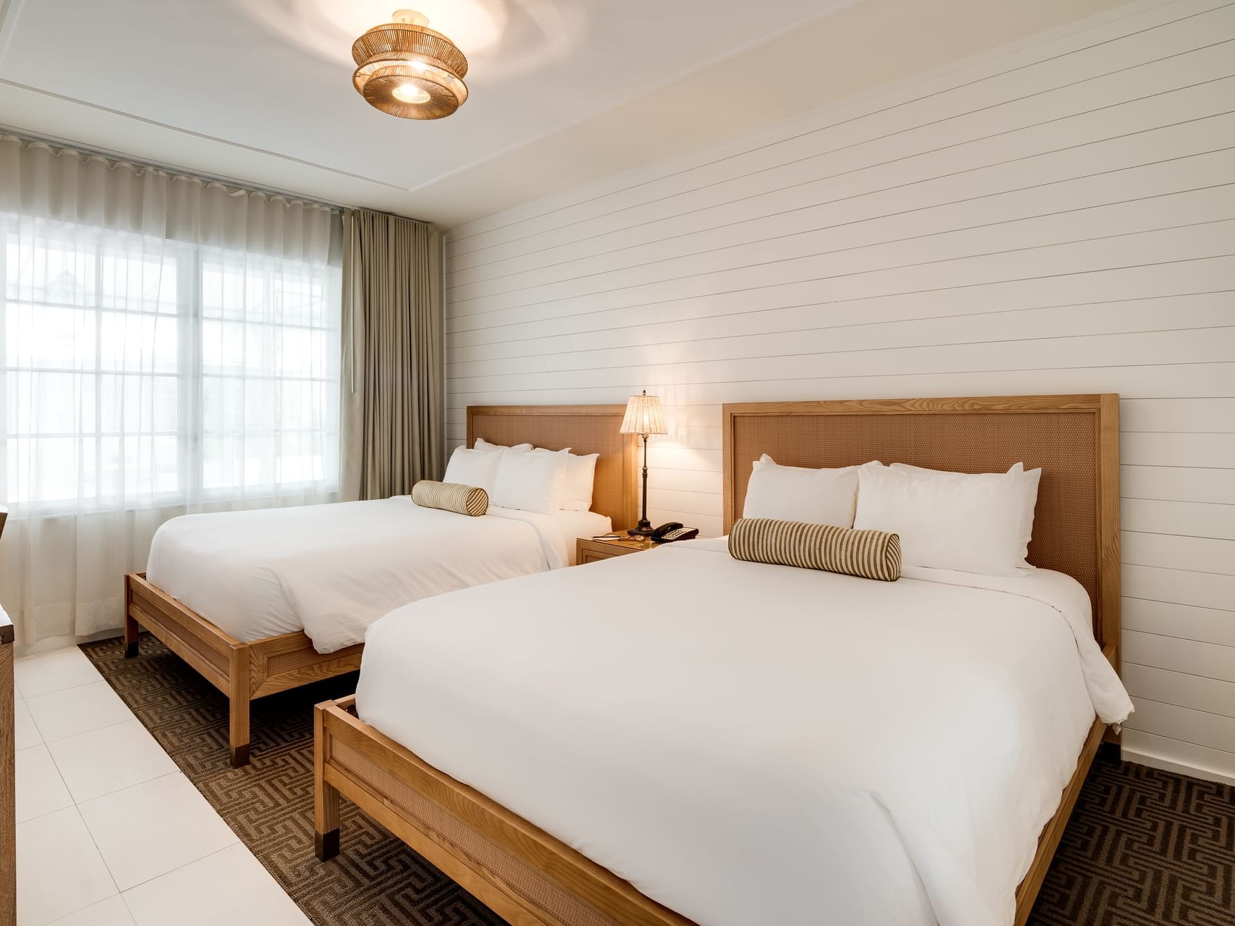 A bedroom with two beds, wooden headboards, white linens, pillows, and a lit lamp on a nightstand.