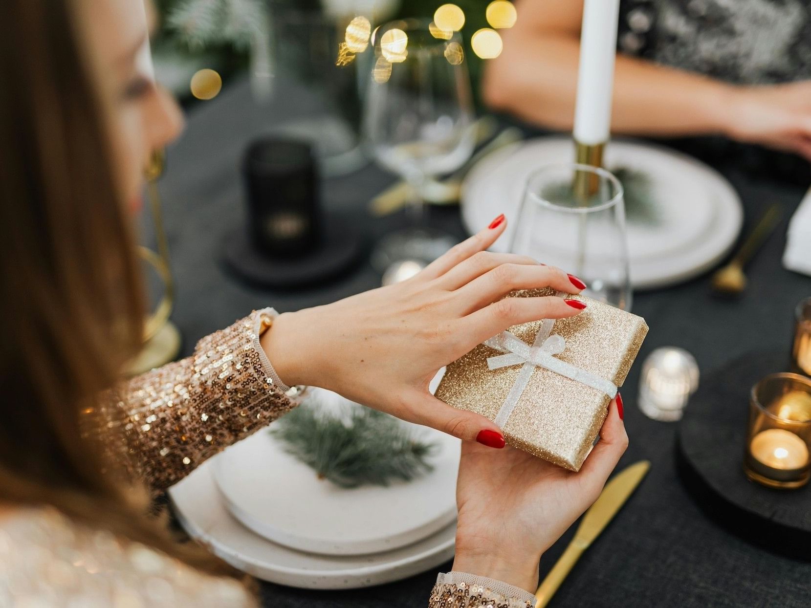 Woman with gold sequined sleeve and red nails holds gold wrapped gift with white ribbon at a dinner table.