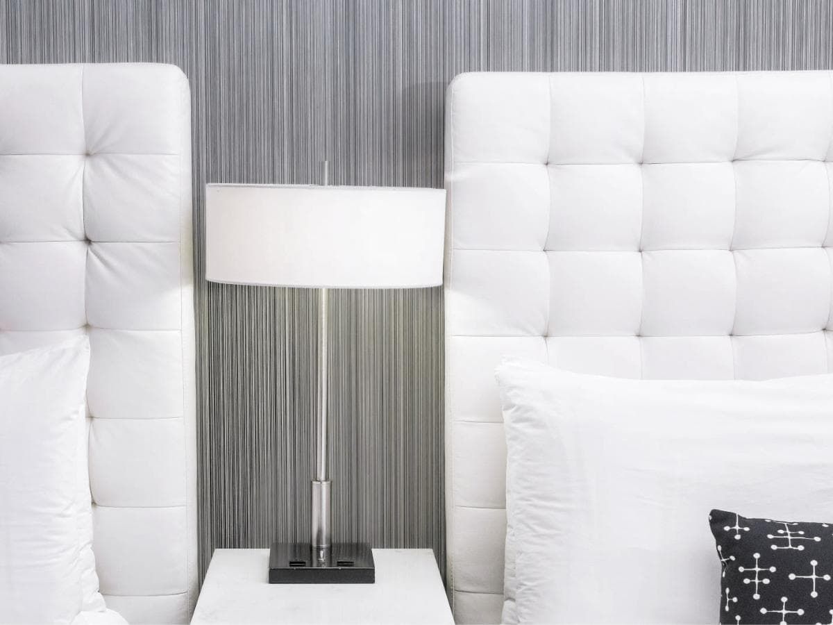 A close-up of a hotel bed with a tufted headboard, white bedding, and a lamp on a nightstand.