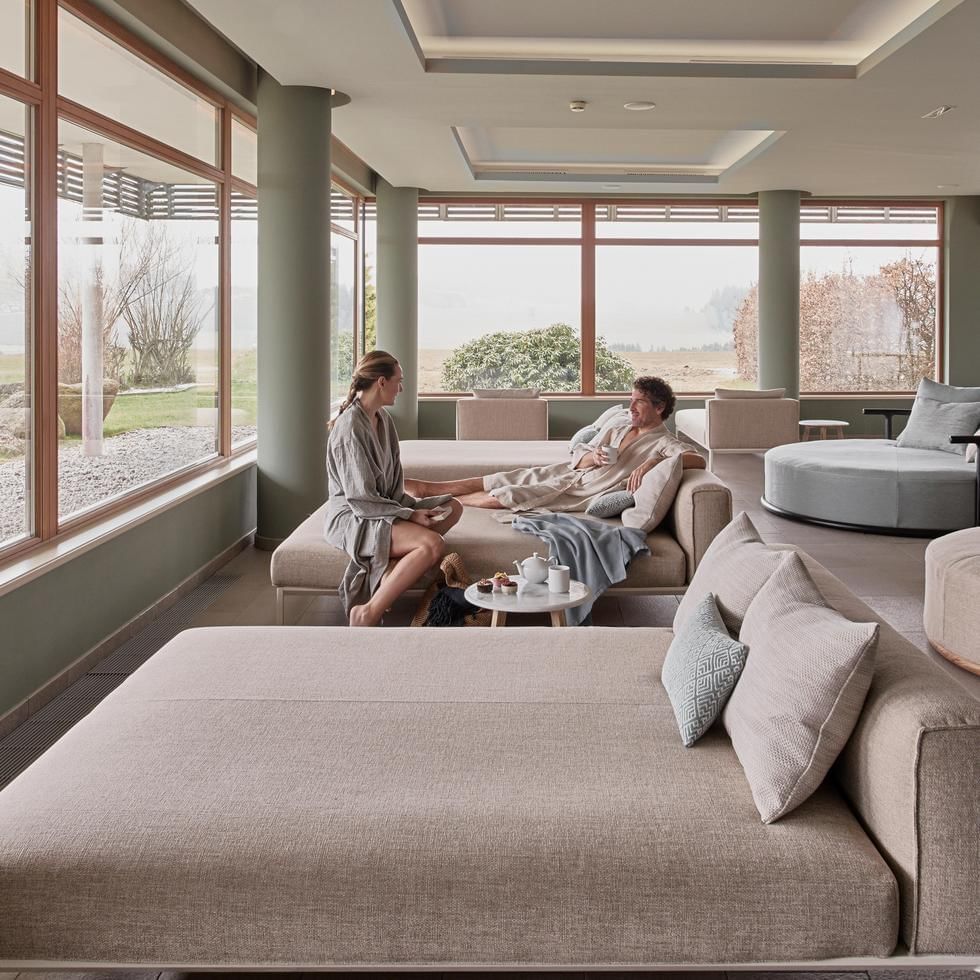 Due persone si rilassano su un divano in una sala con grandi finestre e vista sul giardino.