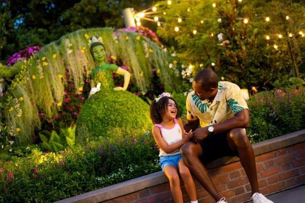 A young girl in a tiara and a man sit on a ledge laughing in front of a topiary shaped like Princess Tiana at EPCOT's International Flower and Garden Festival.