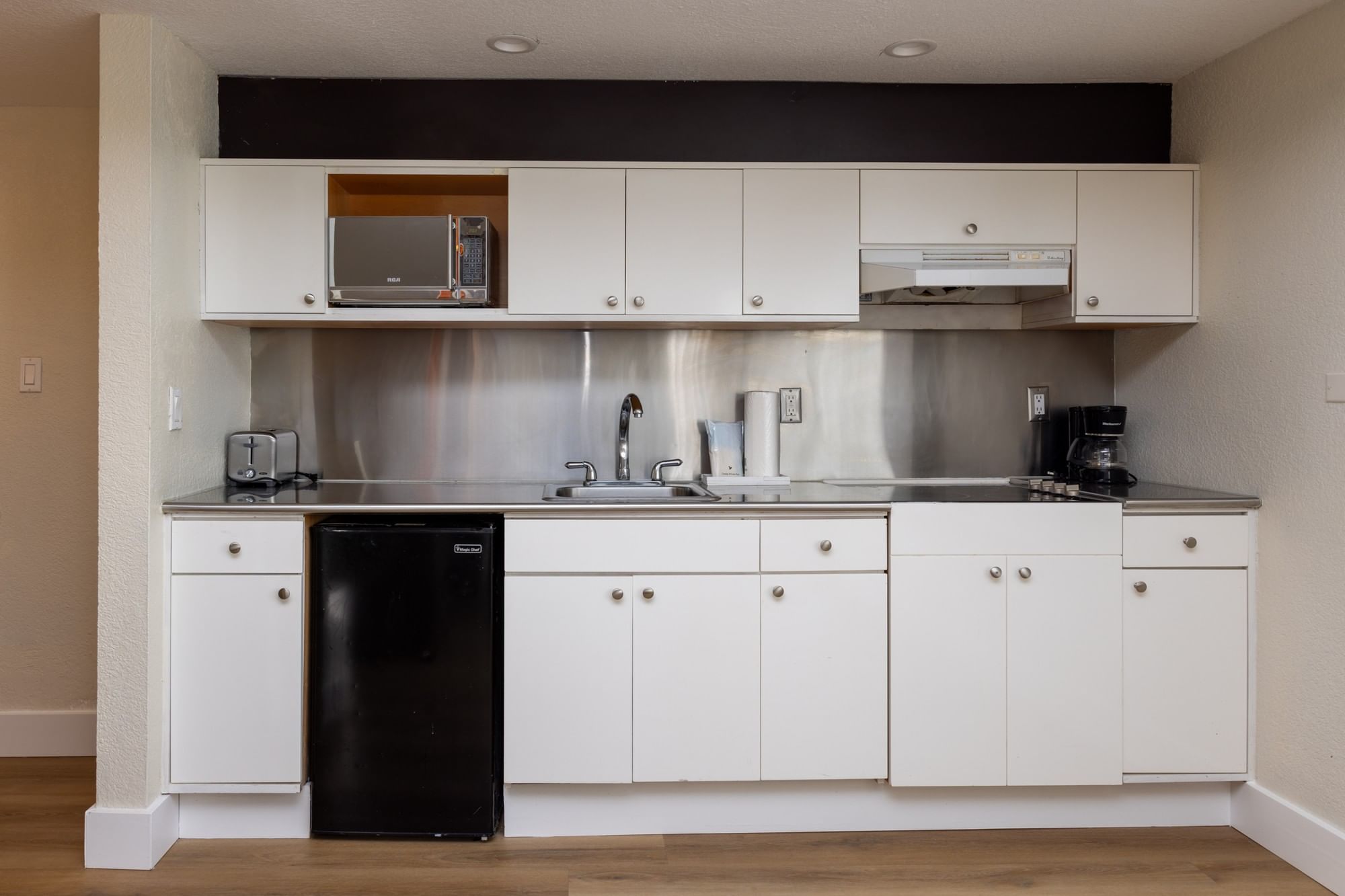 A compact kitchenette with white cabinets, stainless steel backsplash, microwave, coffee maker, sink, and mini fridge.