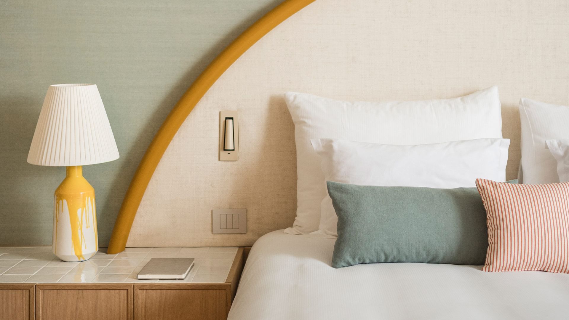 A cozy bed with striped pillows and a yellow lamp on a tiled nightstand.