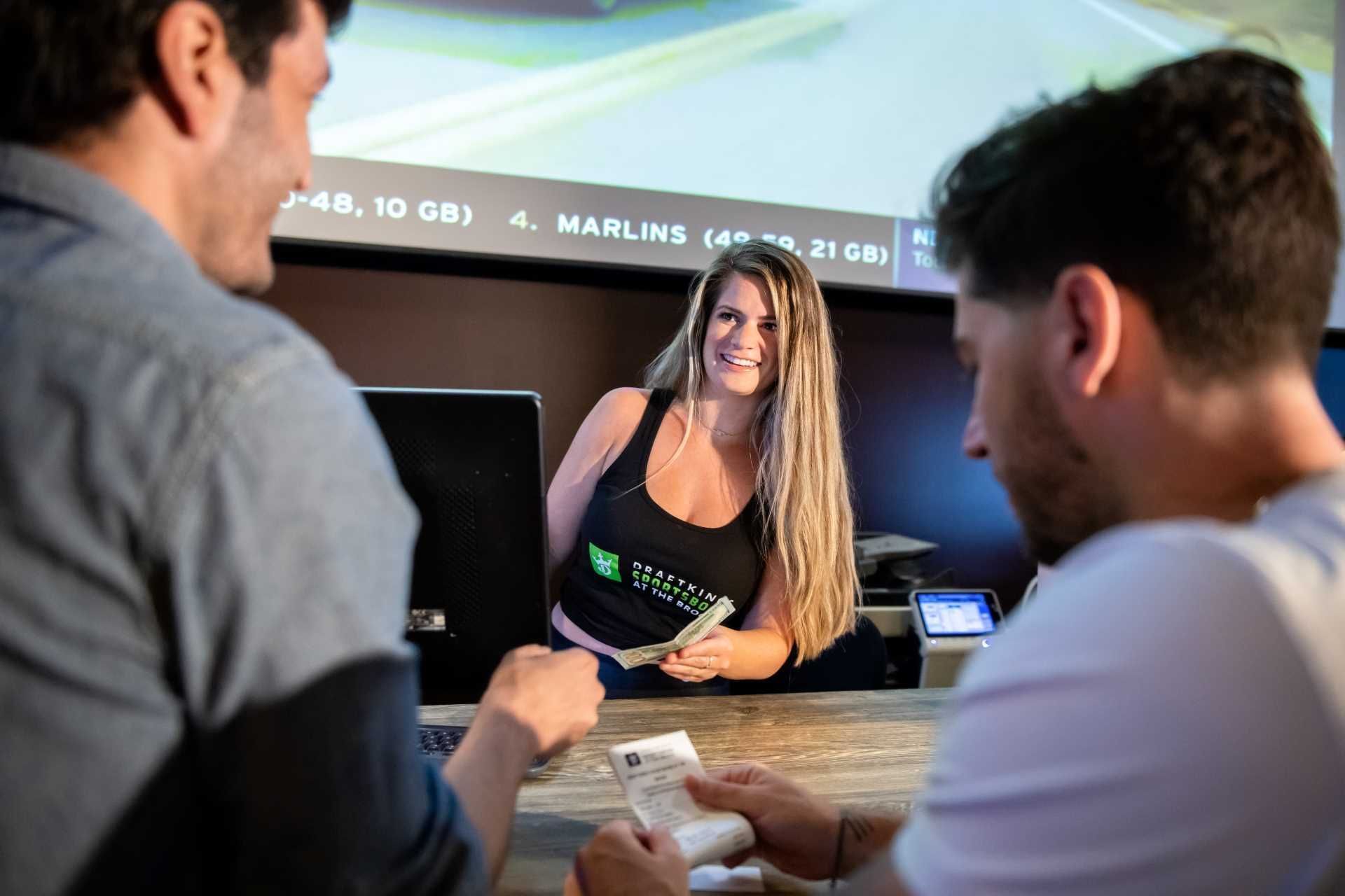 DraftKings Sportsbook attendant serves guests by a betting counter beneath a large sports screen at The Brook