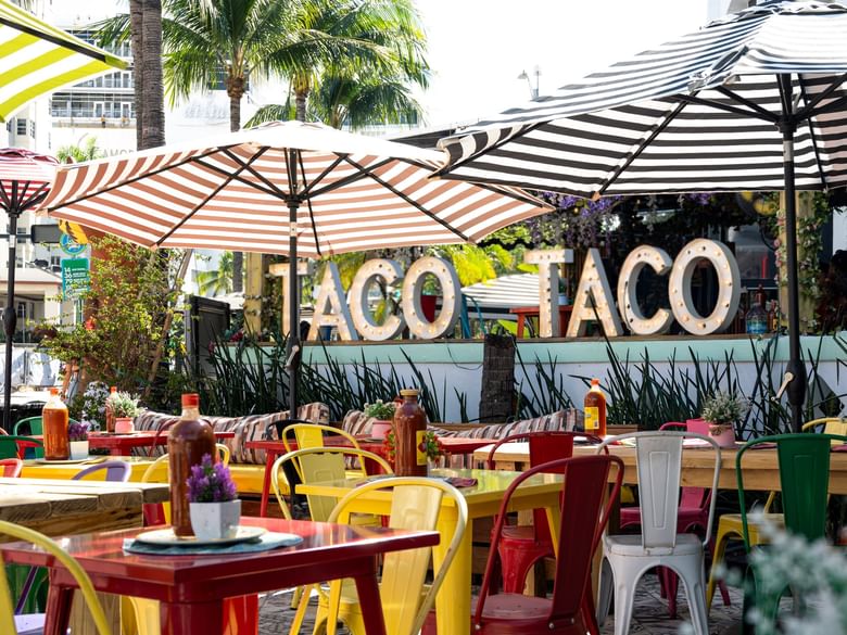 A colorful outdoor dining area with tables, chairs, and umbrellas, and a sign reading 'TACO TACO'.