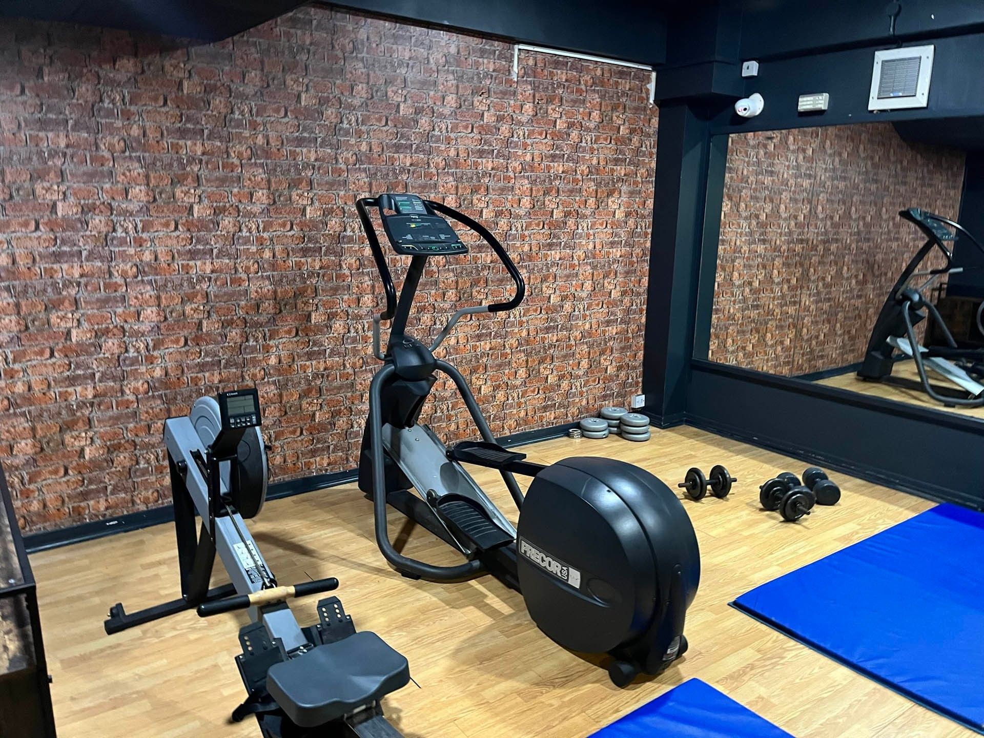 Gym with exercise bike, elliptical trainer, weights, and blue mats on wooden floor, brick wall, and mirror.