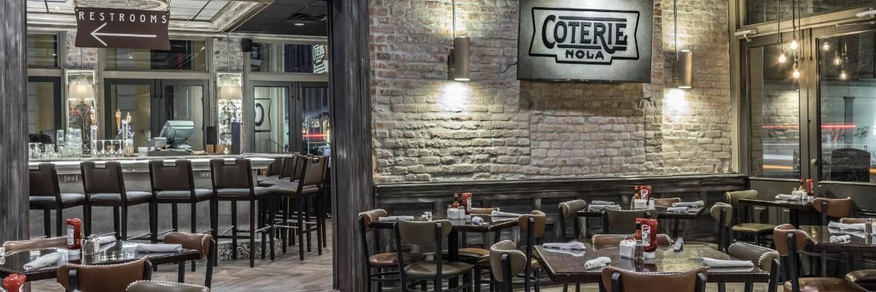 A dining area with tables and chairs, a brick wall, and a sign reading 'Coterie NOLA'.