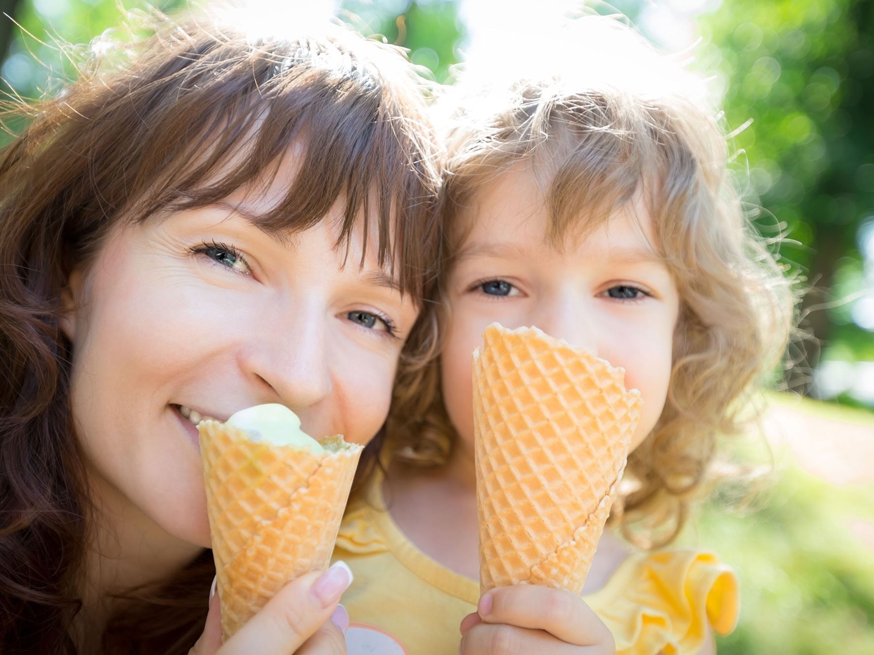 A woman and a child are eating ice cream cones in an outdoor setting with trees and grass in the background.