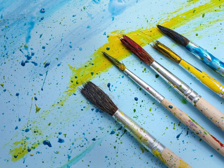 Five used paintbrushes with colorful bristles lying on a blue background with yellow paint splatters at Camino Real