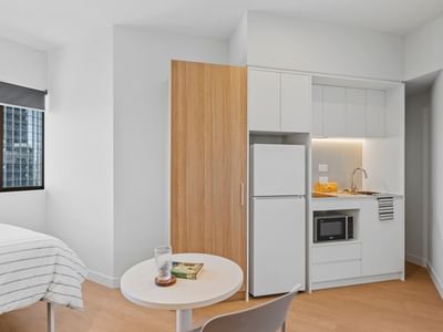 Interior of a clean white room with bed, table, fridge, and kitchen at UniLodge Brisbane City.