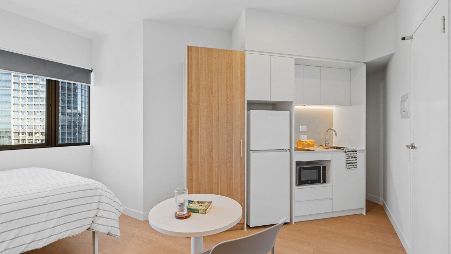 Interior of a clean white room with bed, table, fridge, and kitchen at UniLodge Brisbane City.