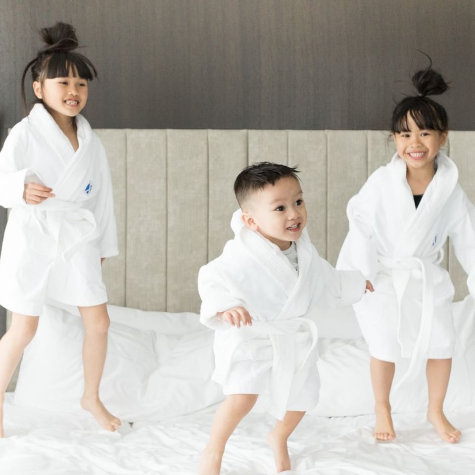 Children in white robes jumping on a bed by a gray tufted headboard under soft lighting at Hotel X Toronto
